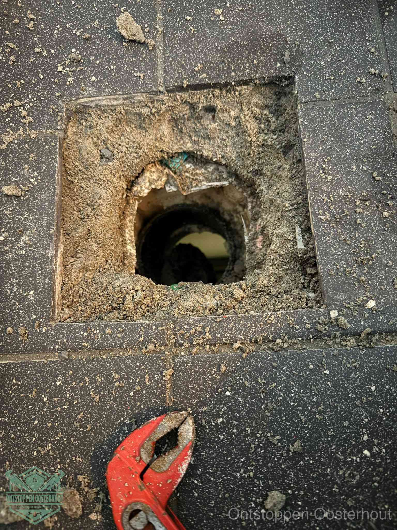 Een geopend rioolputje in een betontegelvloer met losgemaakt rooster en een zichtbare ronde afvoerbuis, het achterhalen waar de afvoerverstopping zit in Oosterhout
