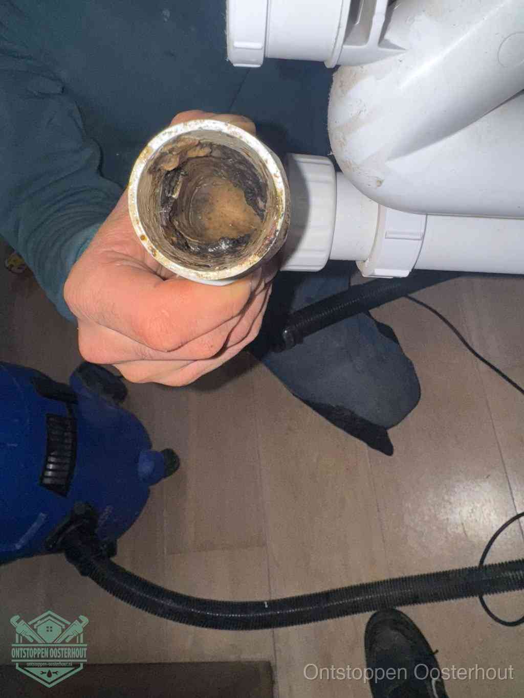 Een hand houdt een losgekoppeld PVC-afvoerbuisstuk vast dat van binnen volledig is dichtgeslibd met vet, vuil en aanslag, de meest voorkomende oorzaak van een verstopte afvoer in Oosterhout